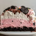 Homemade ice cream cake with layers of chocolate ice cream, hot fudge, Oreo cookie crumbs, strawberry ice cream, and whipped cream, topped with colorful sprinkles.